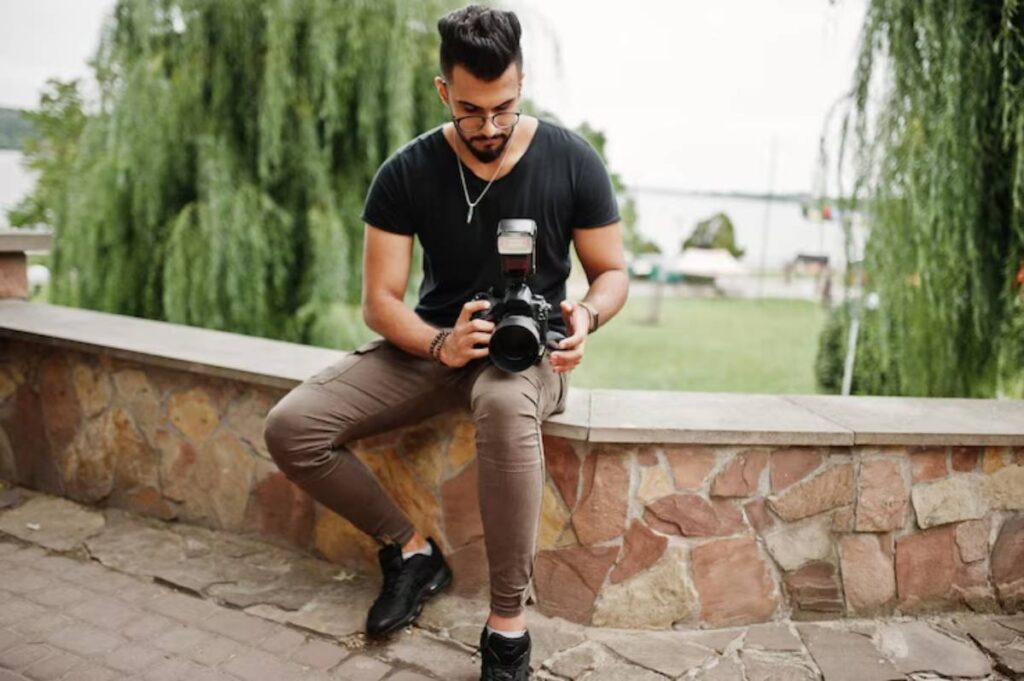 A young man sits on a stone wall with a camera in hand, surrounded by lush greenery and a scenic backdrop.