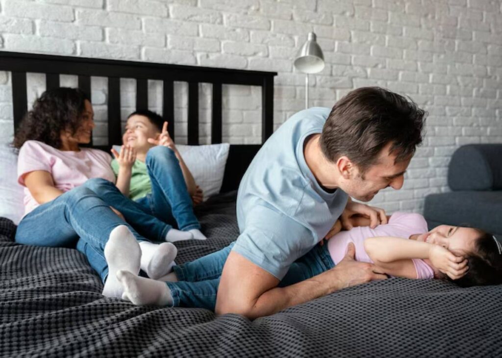 A playful father leans over a child on a bed, while two other children sit nearby, engaged in a fun family moment.