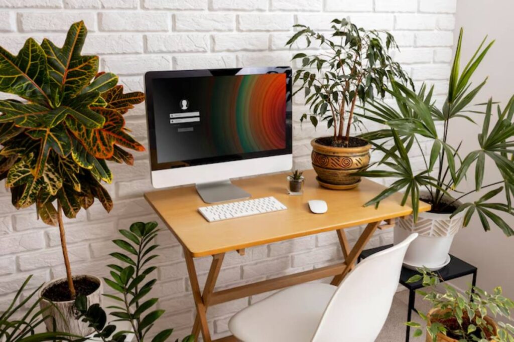 A stylish home office setup featuring a computer, a wooden desk, and various lush green plants against a white brick wall.
