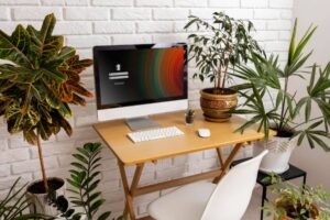 A stylish home office setup featuring a computer, a wooden desk, and various lush green plants against a white brick wall.