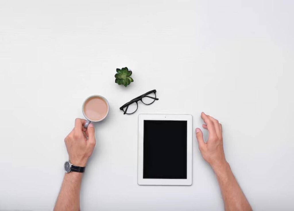 Aerial view of a workspace with a hand holding a coffee cup, an empty tablet, glasses, and a small succulent plant on a white surface.