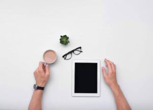 Aerial view of a workspace with a hand holding a coffee cup, an empty tablet, glasses, and a small succulent plant on a white surface.