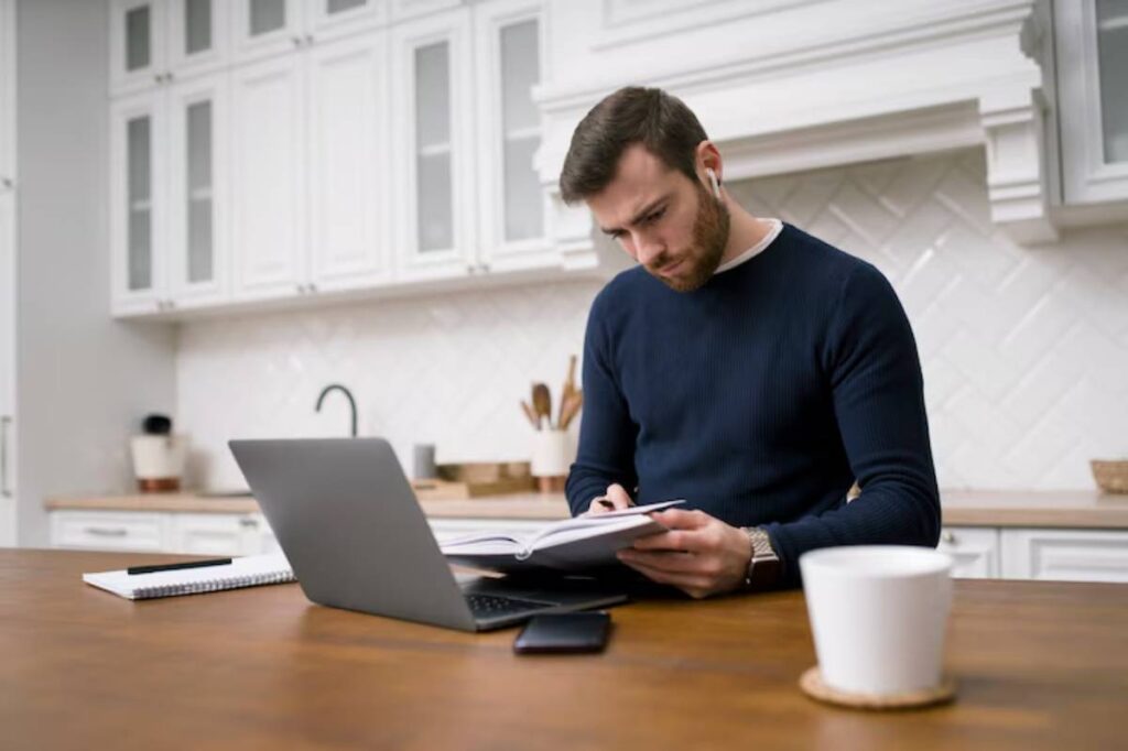 A person wearing a navy sweater is seated at a wooden table, reviewing notes with a laptop, notebook, and coffee cup nearby.