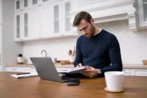A person wearing a navy sweater is seated at a wooden table, reviewing notes with a laptop, notebook, and coffee cup nearby.