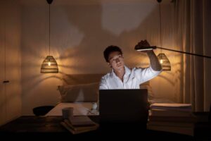 A person in a white shirt is working intently on a laptop in a dimly lit bedroom with warm pendant lights and books visible.