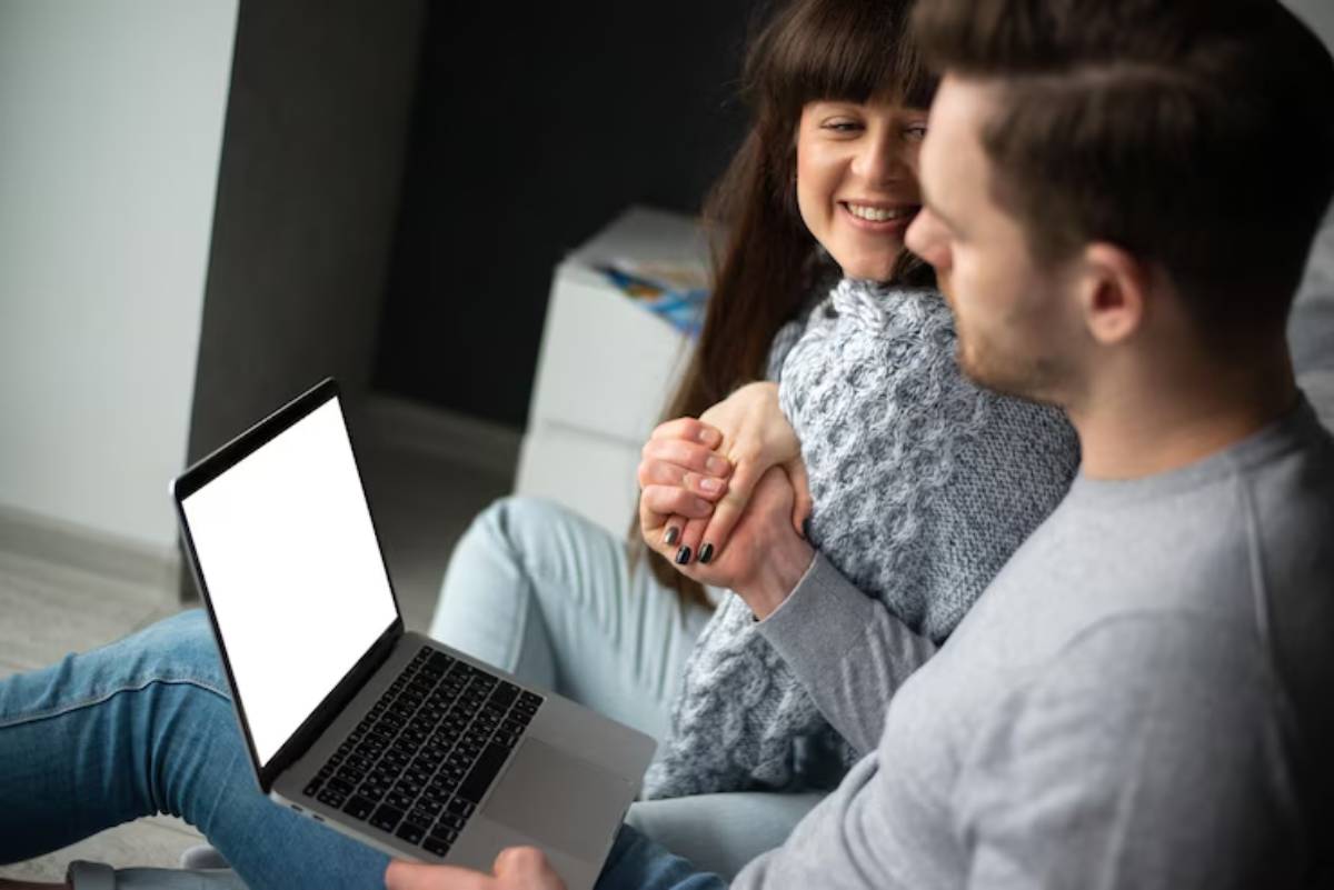 A couple sits closely together, sharing an intimate moment while holding hands, with a laptop in front displaying a blank screen.