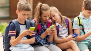 A group of children sitting together, each focused on their smartphones, wearing backpacks, and dressed in casual summer clothing.