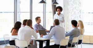 A diverse group of professionals engaged in a meeting at a modern conference table, with a presenter sharing information.