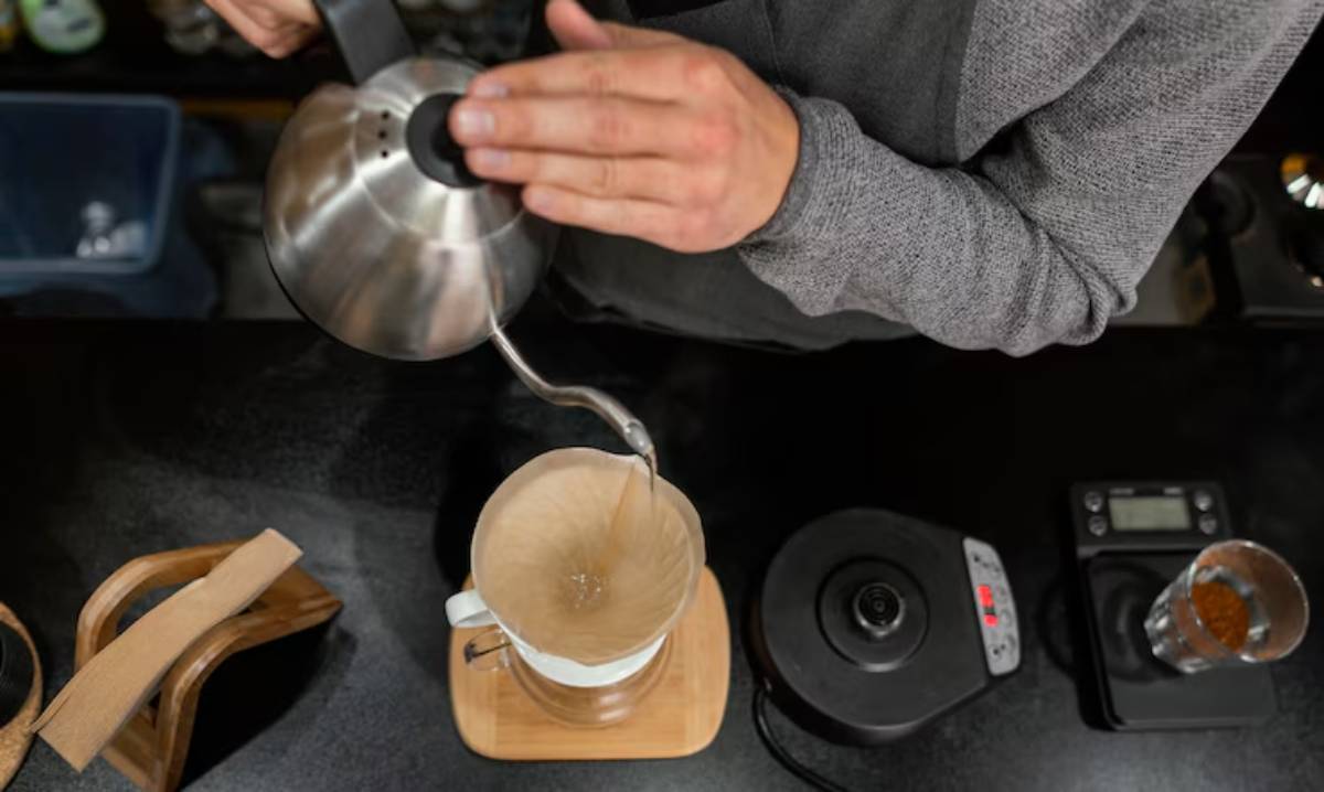 A person pours hot water from a kettle into a coffee filter perched atop a mug, preparing to brew coffee on a black countertop.