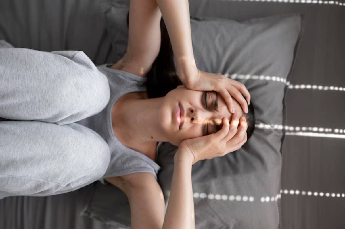 A woman in gray pajamas lies on a bed, cradling her head in pain, with sunlight streaming through blinds casting stripes on her.