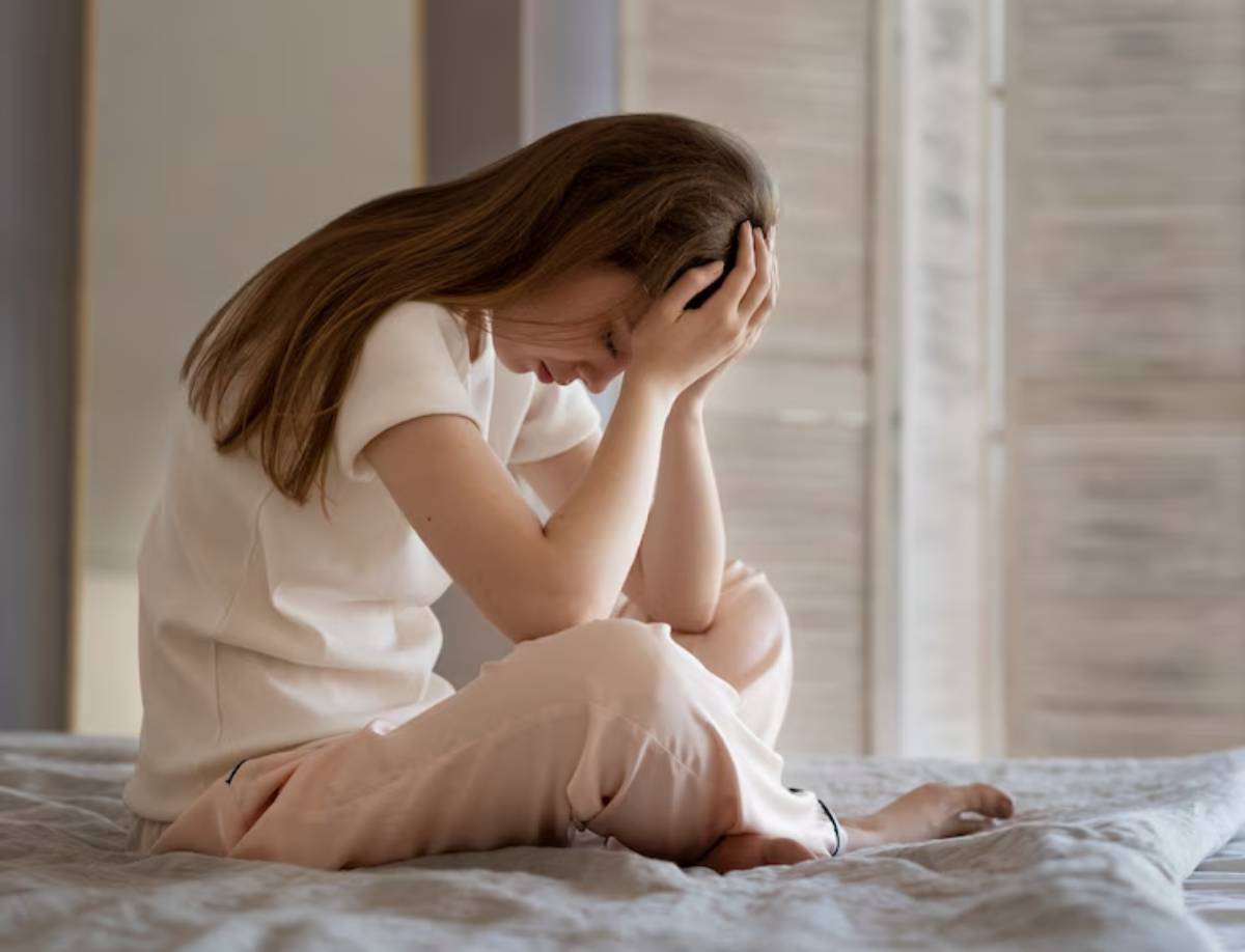 A person sits on a bed, hunched over with their hands covering their face, conveying a sense of distress or contemplation in a serene room.