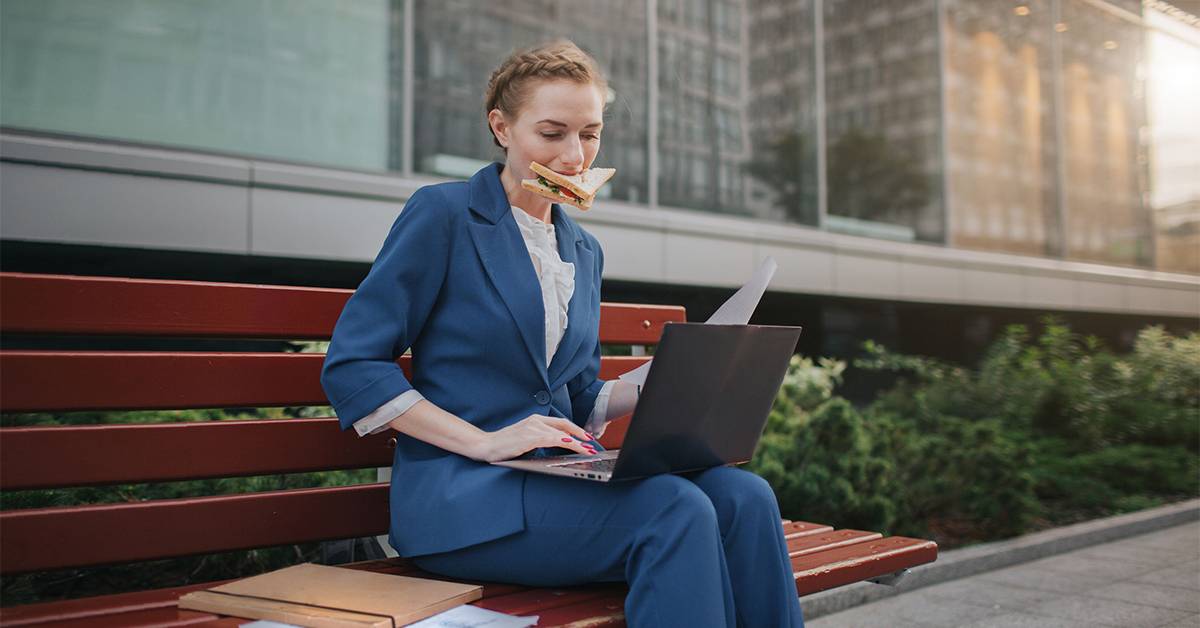 A professional in a blue suit works on a laptop while sitting on a bench outside a modern office building.