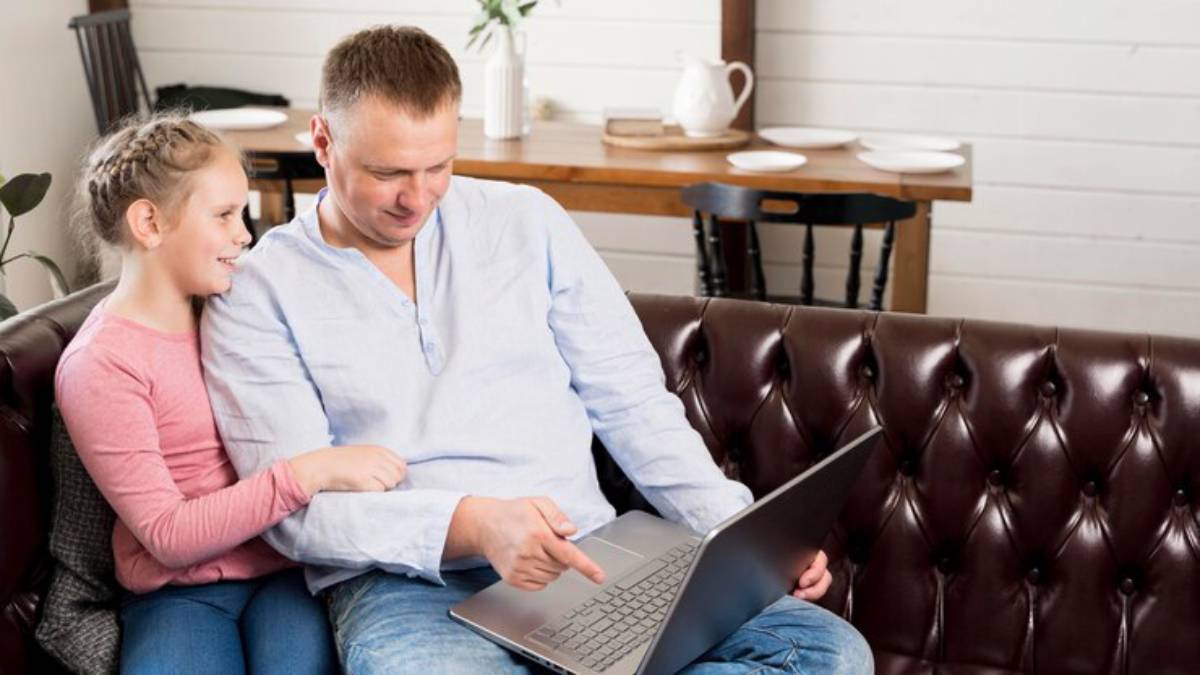 A man interacts with a child on a couch, both focused on a laptop in a cozy, well-lit living space with a wooden table in the background.
