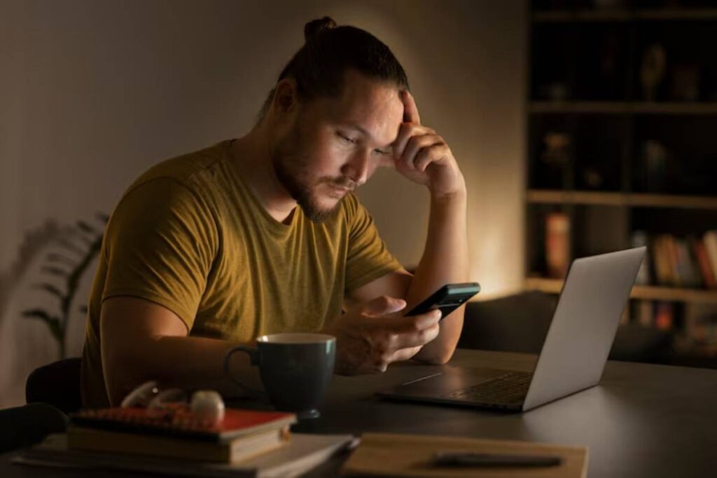A person in a yellow shirt sits at a desk, holding a phone, with a laptop and a cup of coffee nearby, in a dimly lit room.