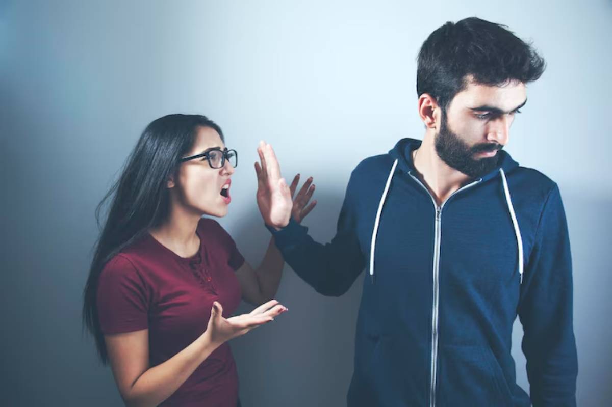 A woman in a burgundy shirt gestures while a man in a blue hoodie turns away, suggesting a tense conversation in a neutral setting.