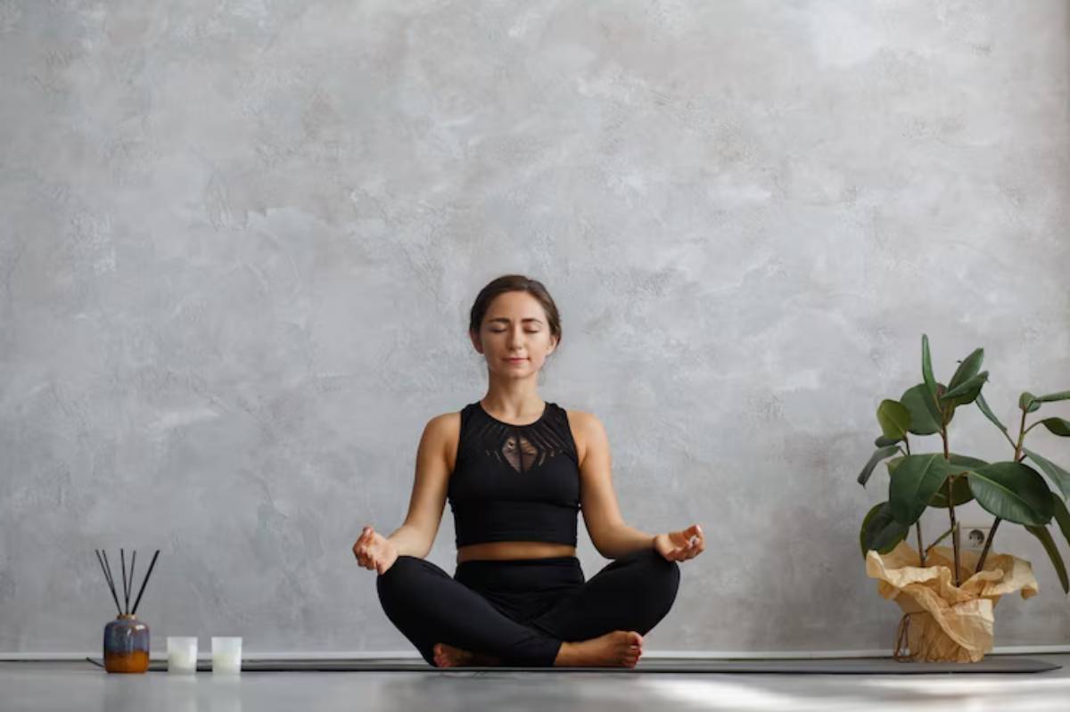 A person in black yoga attire sits in a meditative pose on a mat, surrounded by candles and a potted plant against a grey wall.