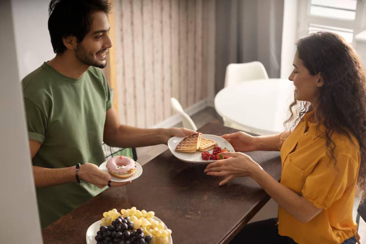 A person hands a plate of waffles and raspberries to another person, while holding a donut with a candle in a cozy kitchen setting. 