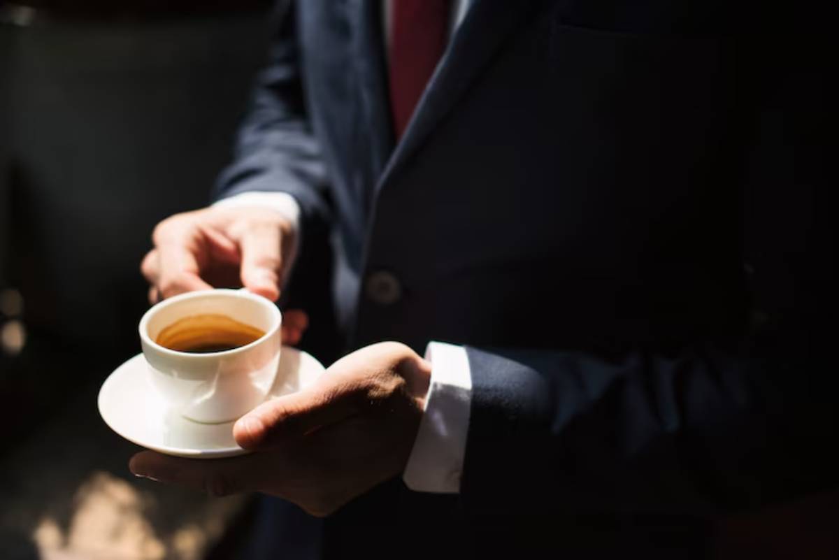 A person in a dark suit holds a small white cup of coffee on a saucer, illuminated by soft light.