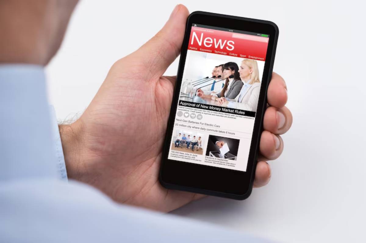 A person holding a smartphone displaying a news website with headlines about new money market rules and electric car batteries.