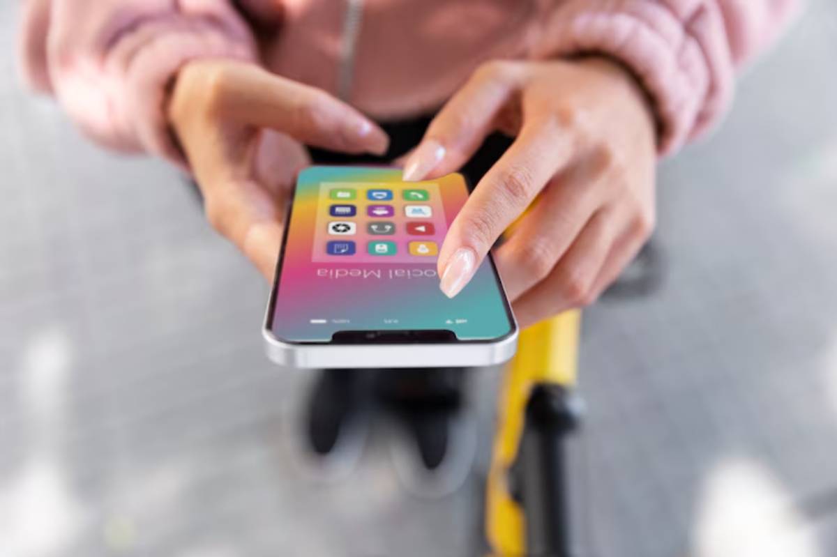 A close-up of a person's hands holding a smartphone displaying colorful app icons, with a blurred background of pavement and a bike.