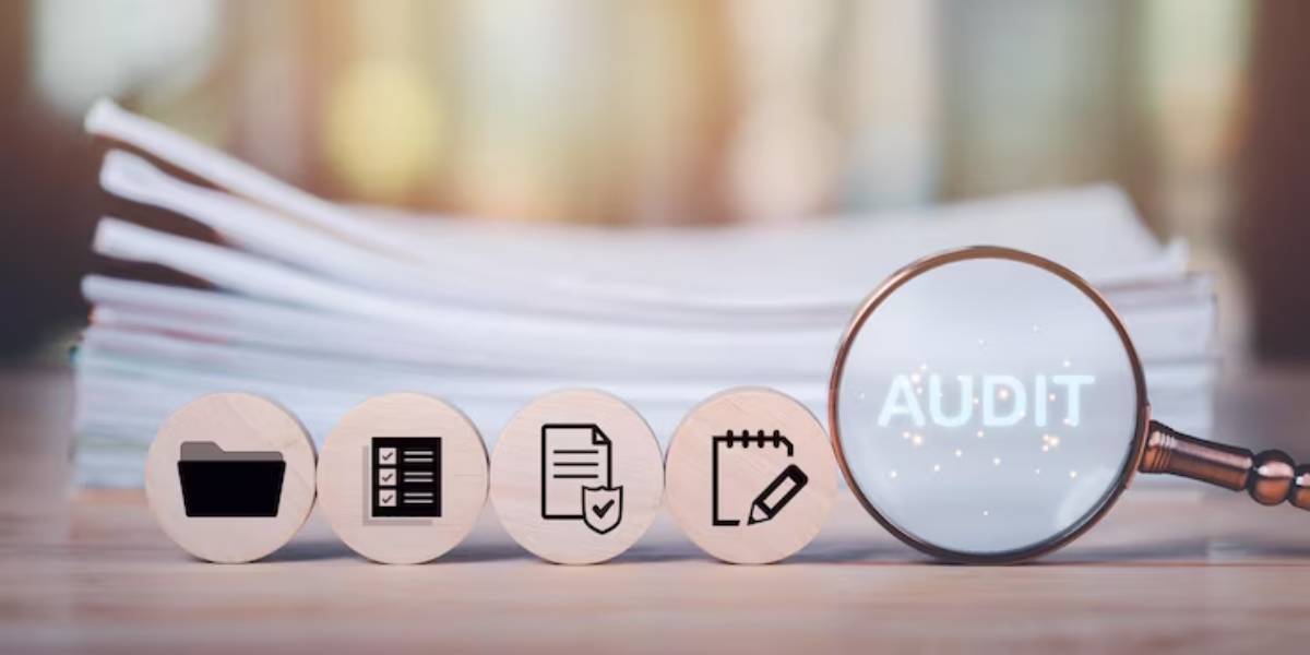 A magnifying glass highlights AUDIT next to wooden icons of a folder, checklist, document, and notebook, with stacked papers in the background.