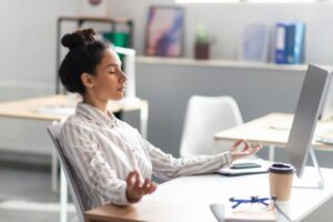 A person sitting at a desk, practicing meditation with hands in a gesture, surrounded by a bright office environment.