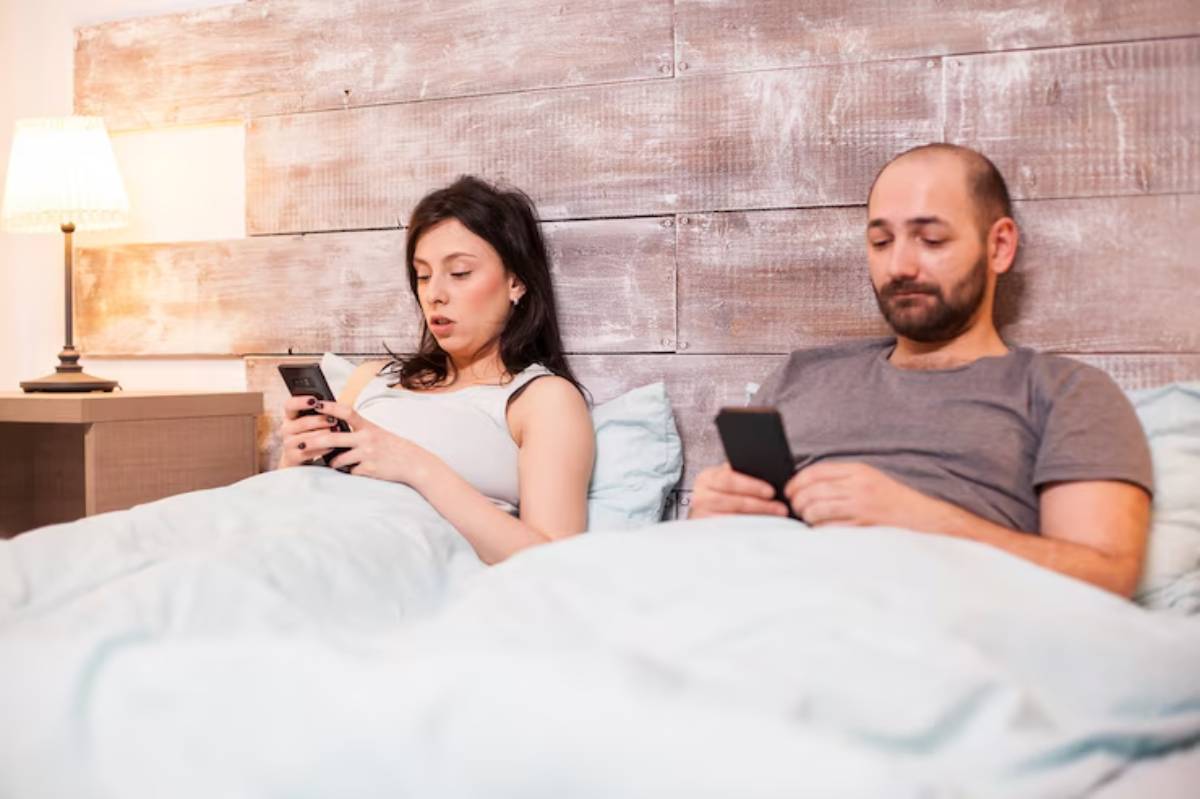 A couple sits in bed, wrapped in blankets, each focused on their smartphones, with a soft lamp illuminating the cozy room.