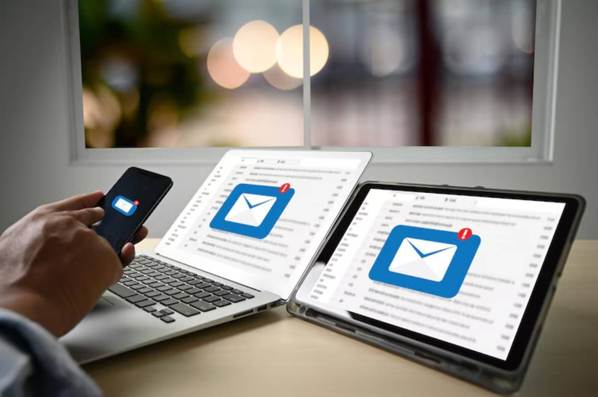 A person checks email notifications on a smartphone, laptop, and tablet, with a bright window in the background.