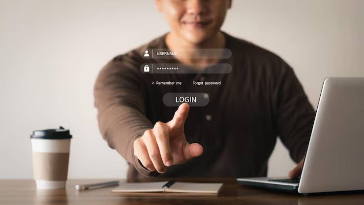 A person points at a login screen on a laptop, with a coffee cup and notebook visible on the table beside them.