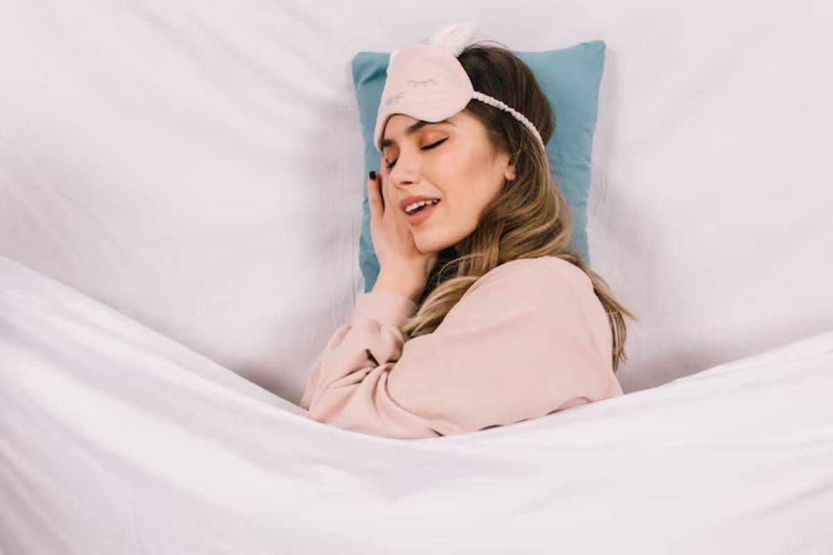 A woman relaxes in bed, wearing a cozy sweatshirt and a soft sleep mask, surrounded by white bedding and a teal pillow.