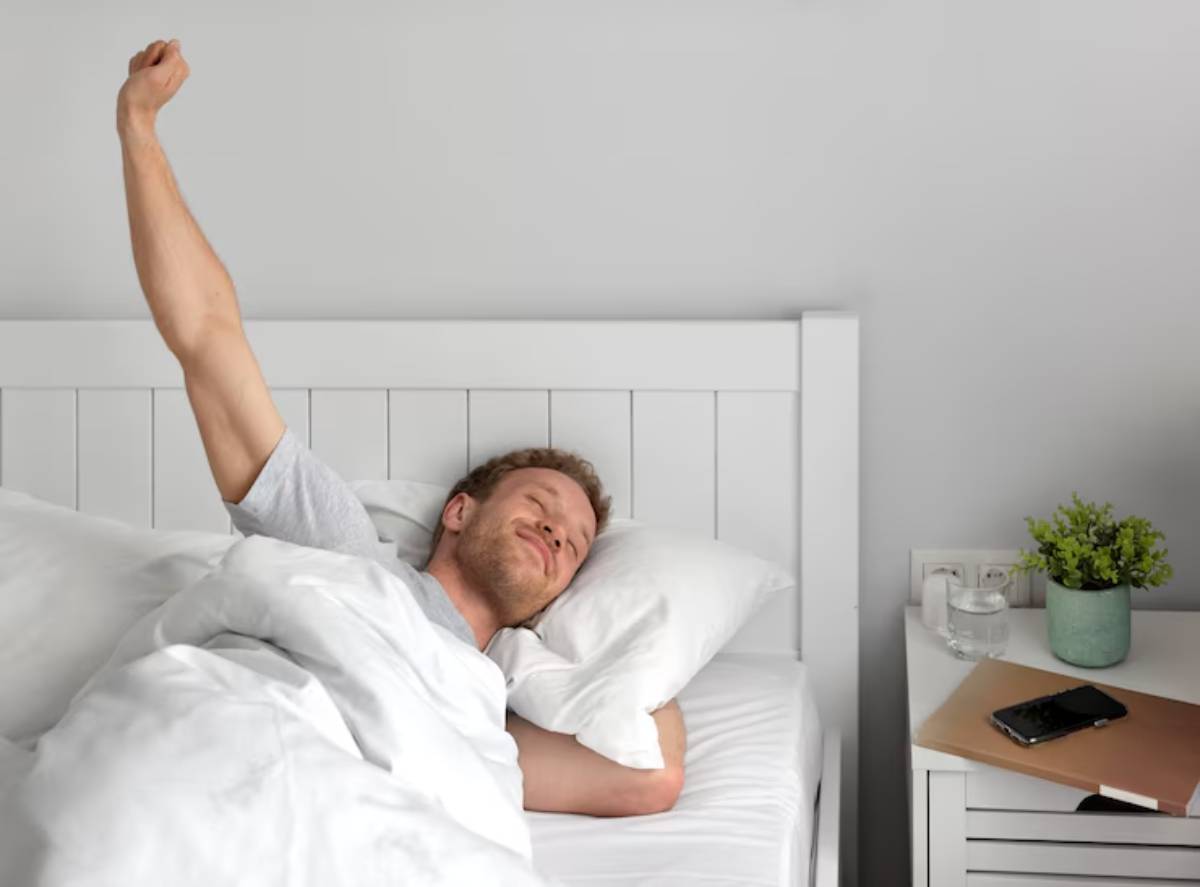  A person stretches in bed, surrounded by white bedding, with a bedside table holding a phone and a potted plant. Natural light fills the room.