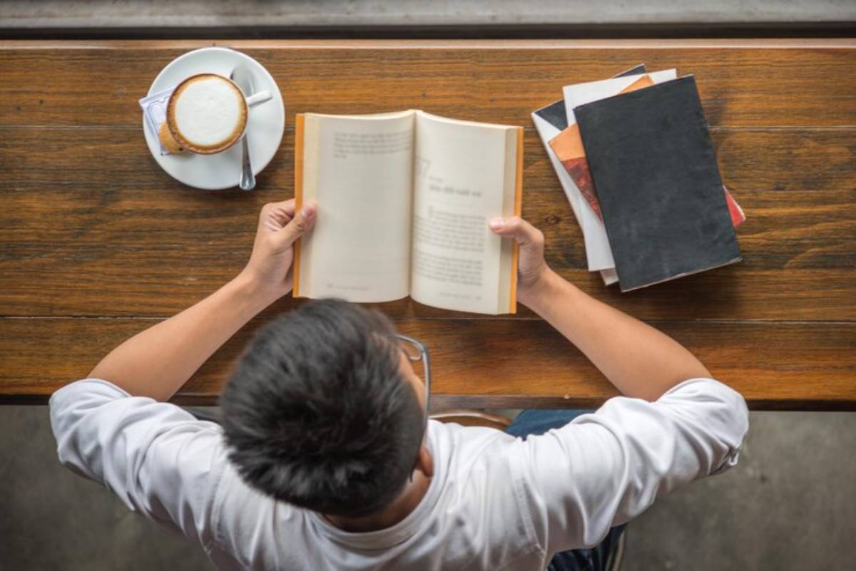 A person reading a book at a wooden table, with a cup of coffee and stacked books nearby, viewed from above.