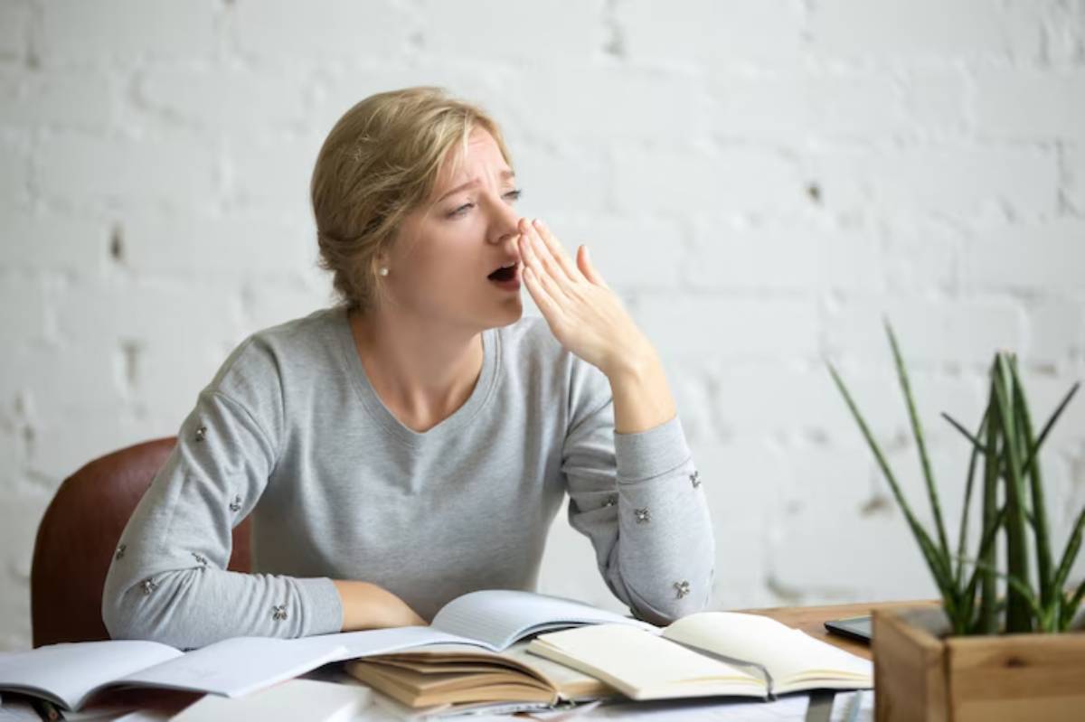 A woman sits at a desk with books and papers, looking contemplative, in a bright, minimalist workspace with a plant in the background. 