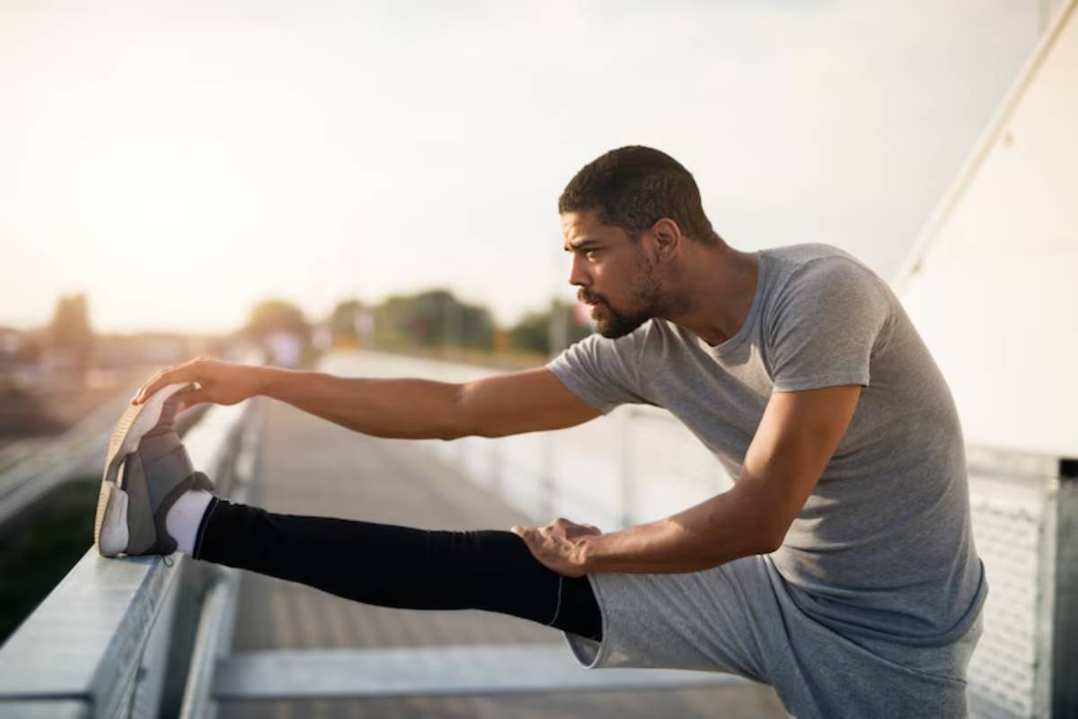 A man in athletic wear stretches his leg on a railing, with a serene outdoor background and soft sunlight illuminating the scene.