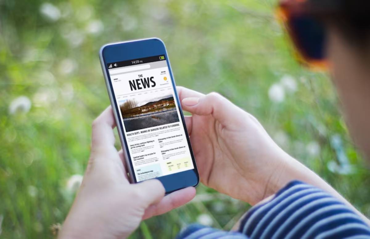 A person reads a news article on a smartphone while sitting outdoors on grass, surrounded by dandelions.