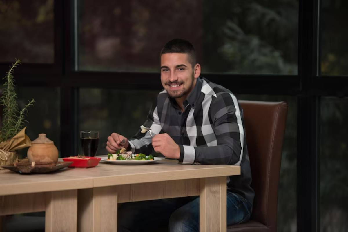 A person sits at a wooden table enjoying a meal, with a glass of dark liquid nearby and salad on a plate. Greenery decorates the scene.