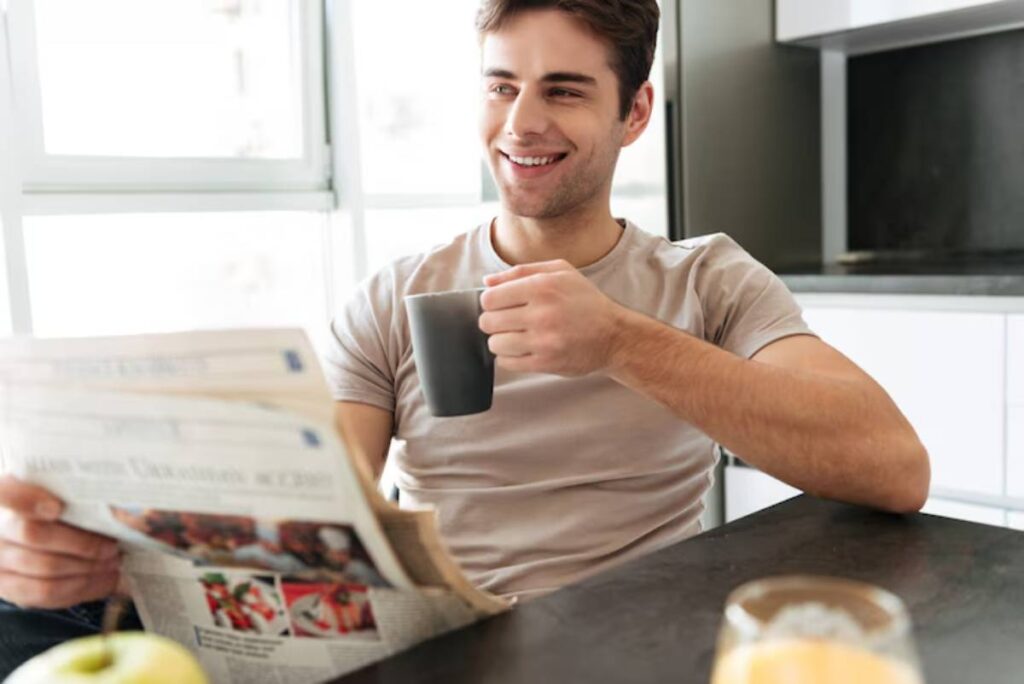 A person sitting at a table holding a coffee mug, reading a newspaper, with an apple and a glass of orange juice nearby.