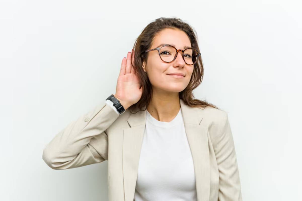 A woman in a beige blazer and white shirt holds her hand to her ear, appearing to listen intently against a plain white background.