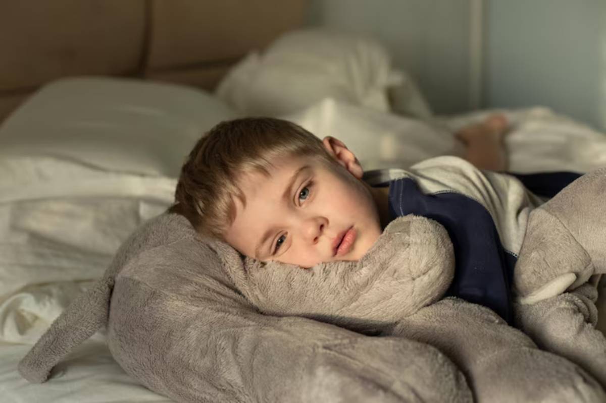 A young child rests peacefully on a bed, cuddling a plush gray animal amidst white sheets and soft blankets.