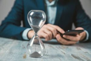 A businessman in a blue suit checks his smartphone as an hourglass sits on the table, symbolizing the passage of time.