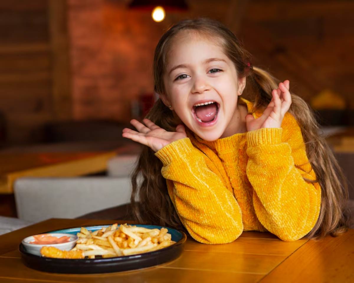 A young girl wearing a cozy yellow sweater sits at a wooden table, resting her chin on her hands near a plate of fries and dipping sauce.