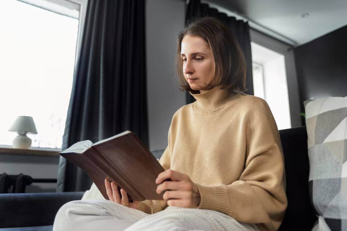 A person wearing a cozy beige sweater sits on a couch, reading a book, with soft natural light streaming through a window.