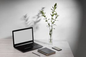 A minimalist workspace featuring a laptop, a notebook, a smartphone, and a vase with greenery, casting gentle shadows on the surface.