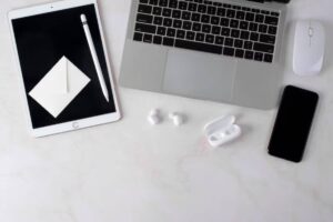 A sleek workspace featuring an iPad, MacBook, smartphone, wireless earbuds, a mouse, and a stylus on a marble surface.