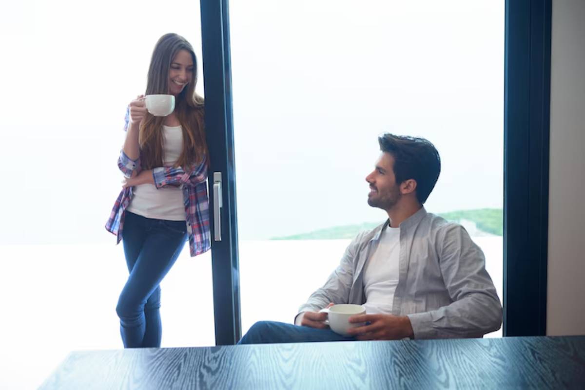 A young woman in a plaid shirt stands by a large window holding a cup, while a young man sits nearby with his own cup.