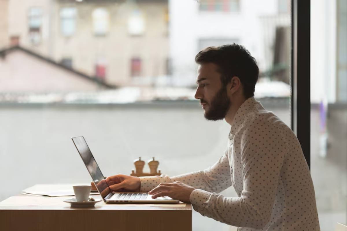 A person in a patterned shirt works on a laptop at a wooden table, with a coffee cup and chess pieces nearby, in a cozy café setting. 