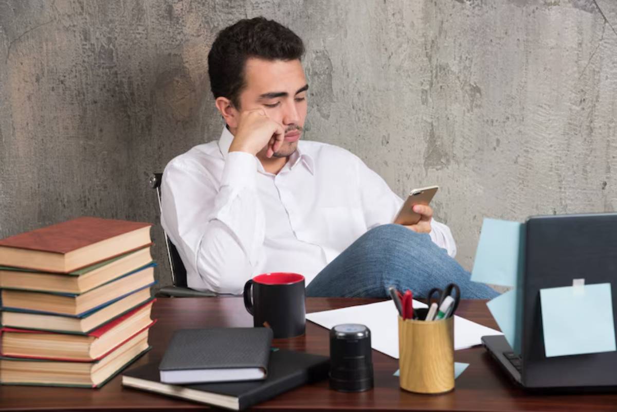 A person sits in an office chair, holding a smartphone, surrounded by a stack of books, a notebook, and a potted plant on a desk.