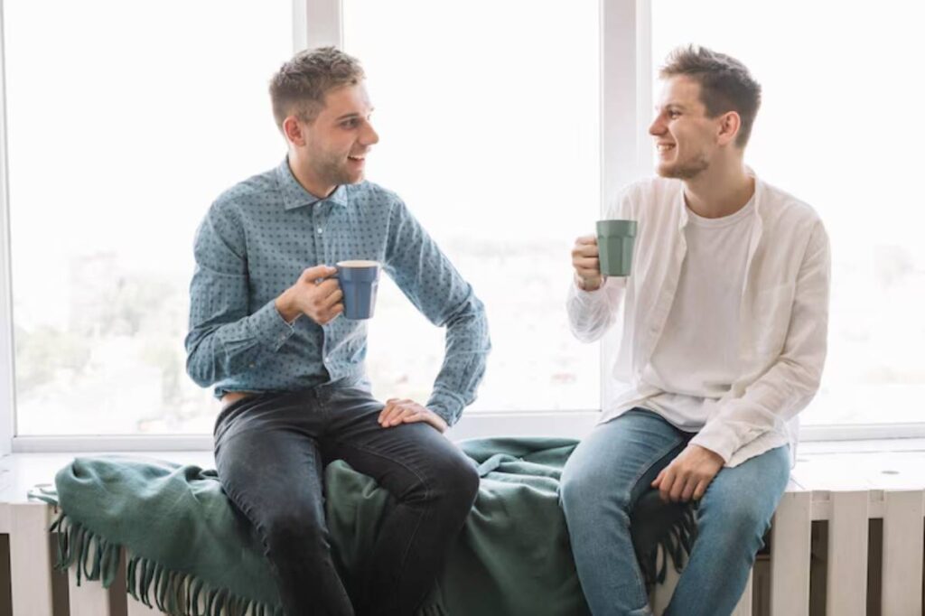 Two individuals seated on a window ledge, enjoying drinks and a cozy atmosphere with a green blanket draped beside them.