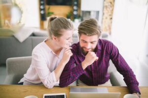 A couple sits closely at a desk, sharing a quiet moment while working on a laptop, surrounded by a cozy, modern living space.