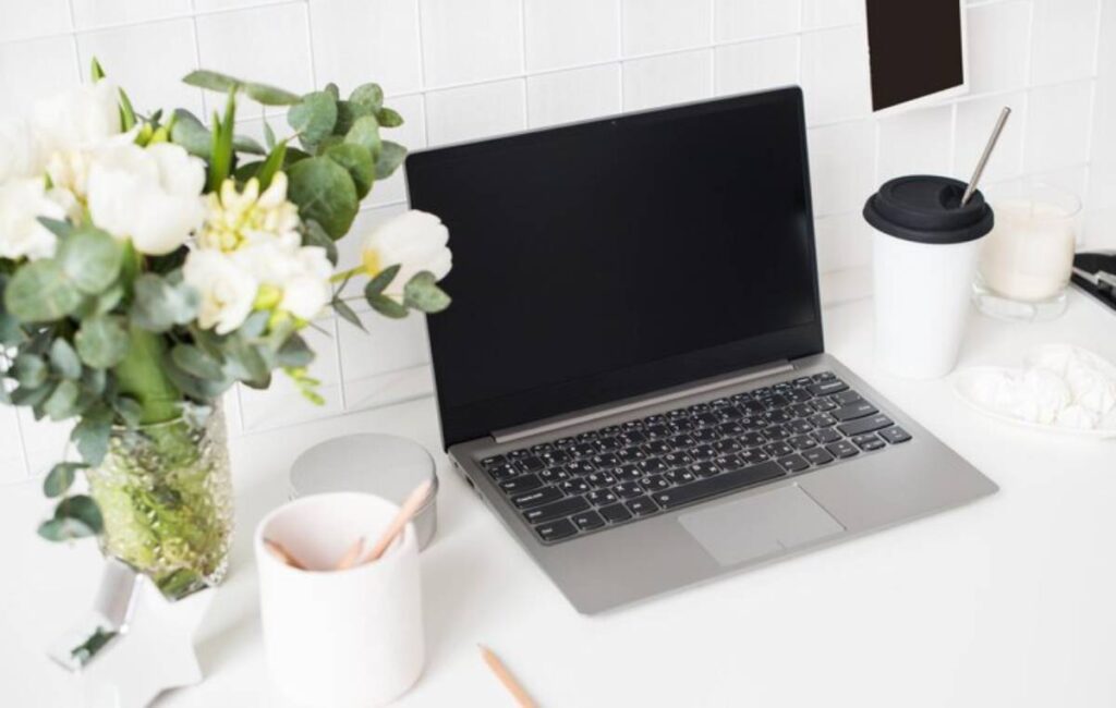 A sleek laptop on a white desk, accompanied by a vase of flowers, a coffee cup, and a small container with utensils.